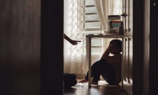 A little Asian boy with a sad and lonely expression hides in the shadows under a table at home, while being scolded by his mother
